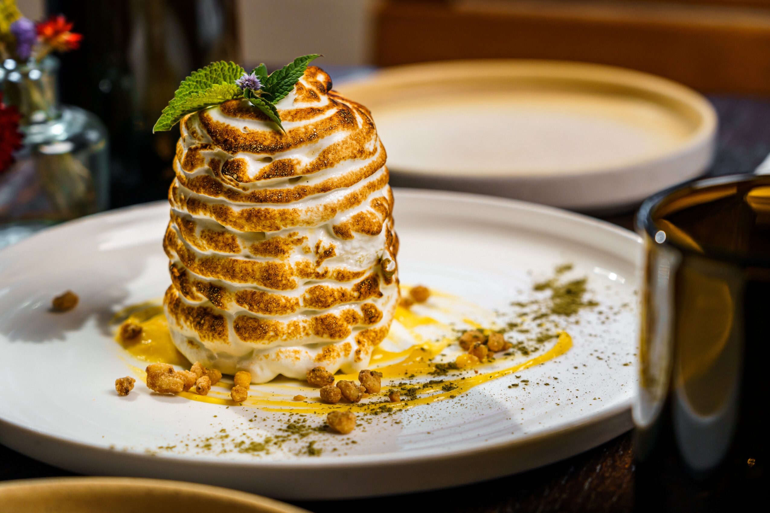 A tall, layered dessert with caramel drizzle, garnished with a mint leaf and purple flower, on a white plate.