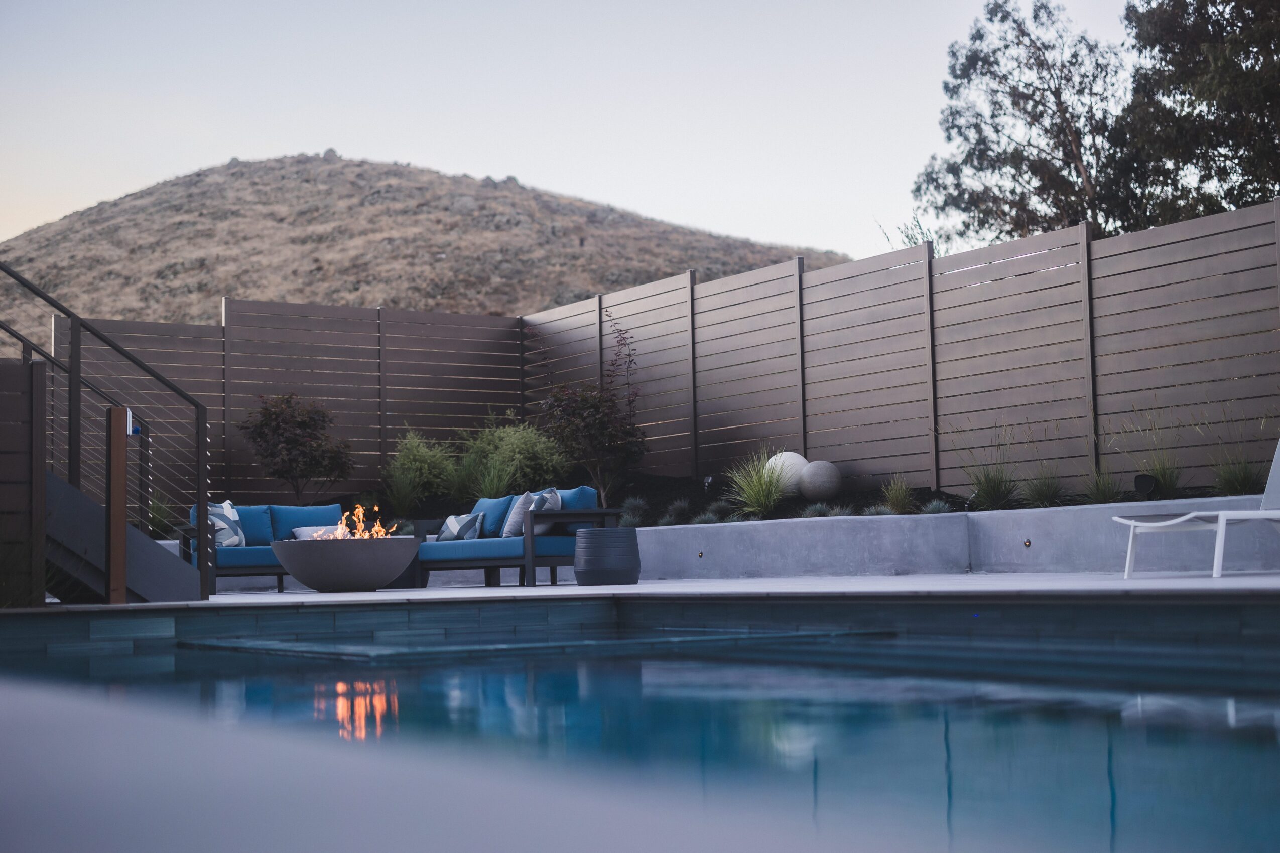 Swimming pool with outdoor seating area, fence, and hillside in background, during daytime.