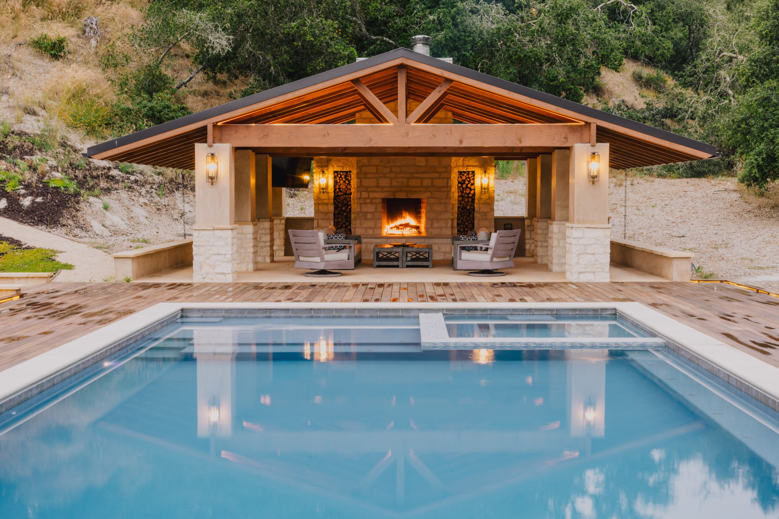 Outdoor pool with a pavilion featuring a fireplace, seating area, and surrounding greenery.