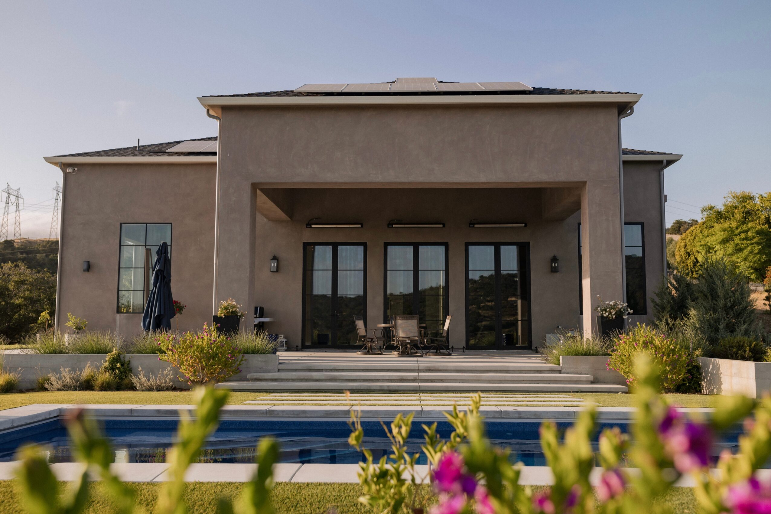 Modern house with large glass doors, outdoor seating, and a swimming pool, surrounded by plants and flowers.