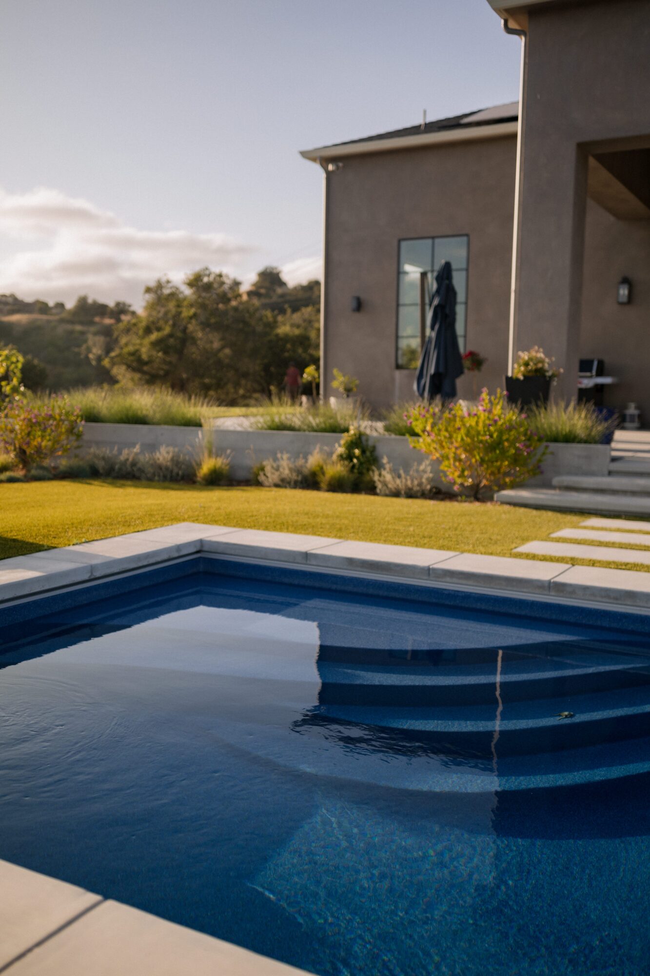House with patio, garden, and swimming pool in backyard, sunny day, trees in background.