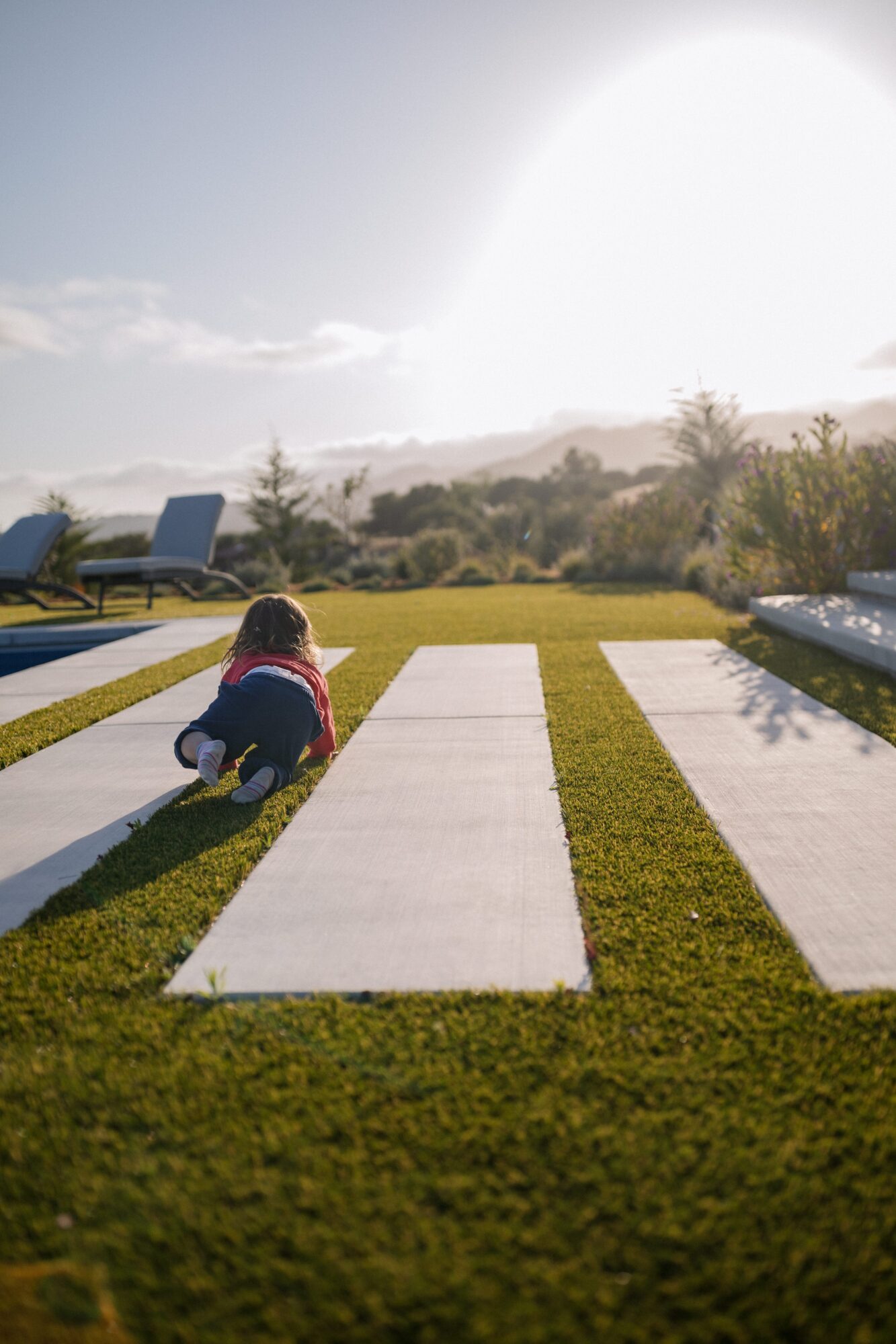 Child crouching on a pathway outdoors with chairs and trees in the background, bright sunlight, and a clear sky.