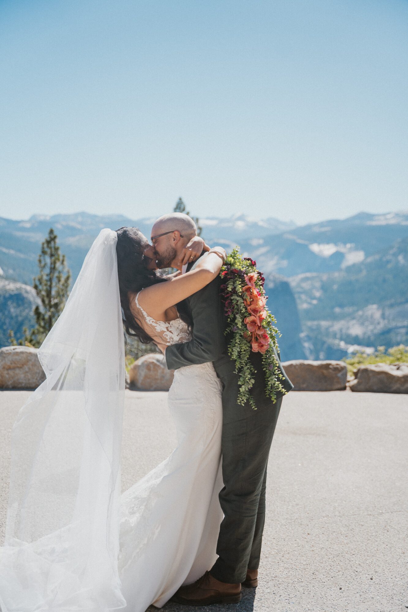 Bride and groom embrace outdoors with mountains and blue sky in background, bride wearing veil and wedding dress, groom in suit, holding a bouquet.