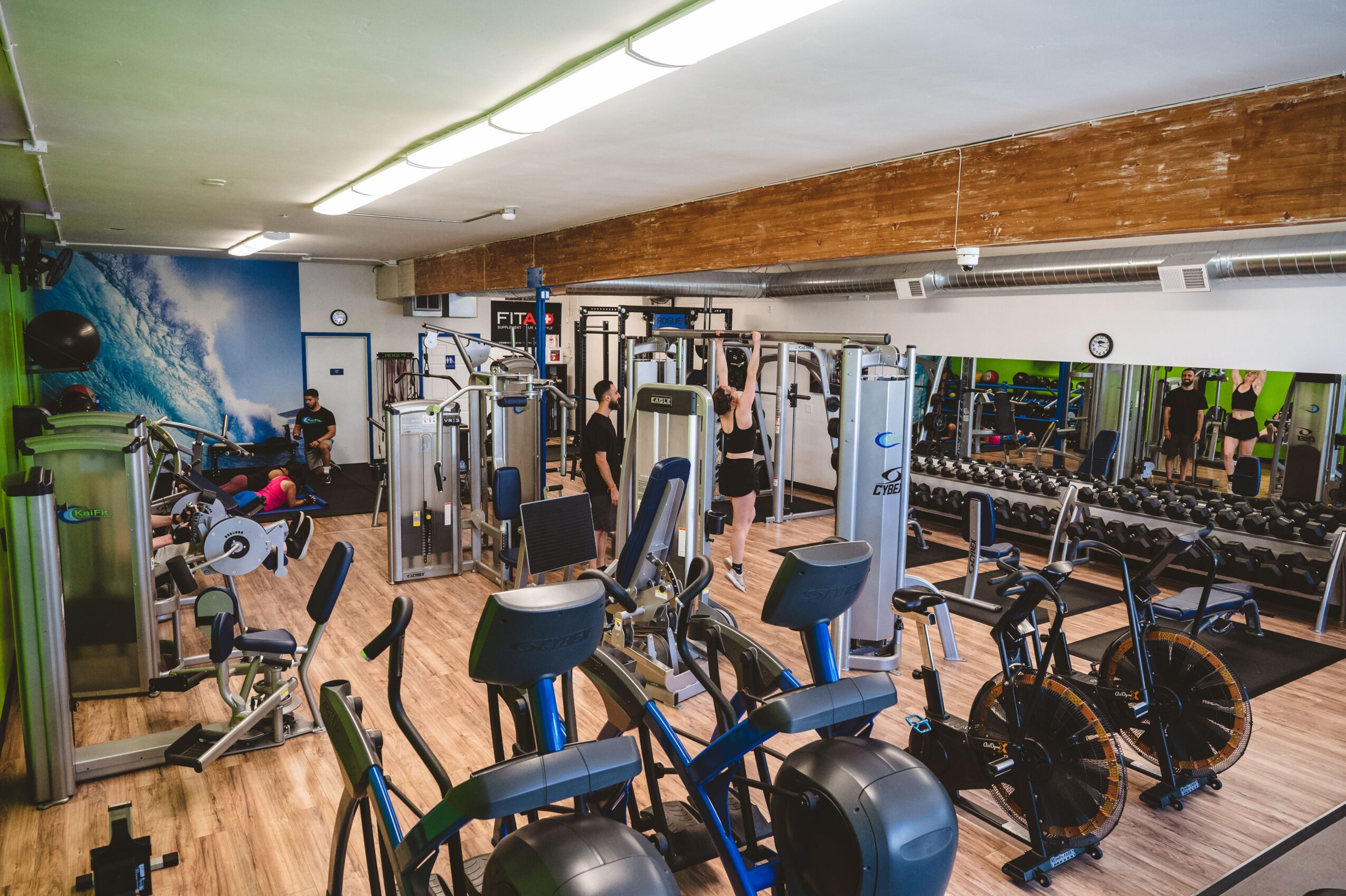 Gym with exercise machines, bikes, and a mirror wall, wooden ceiling, and colorful wall mural, well-lit space.