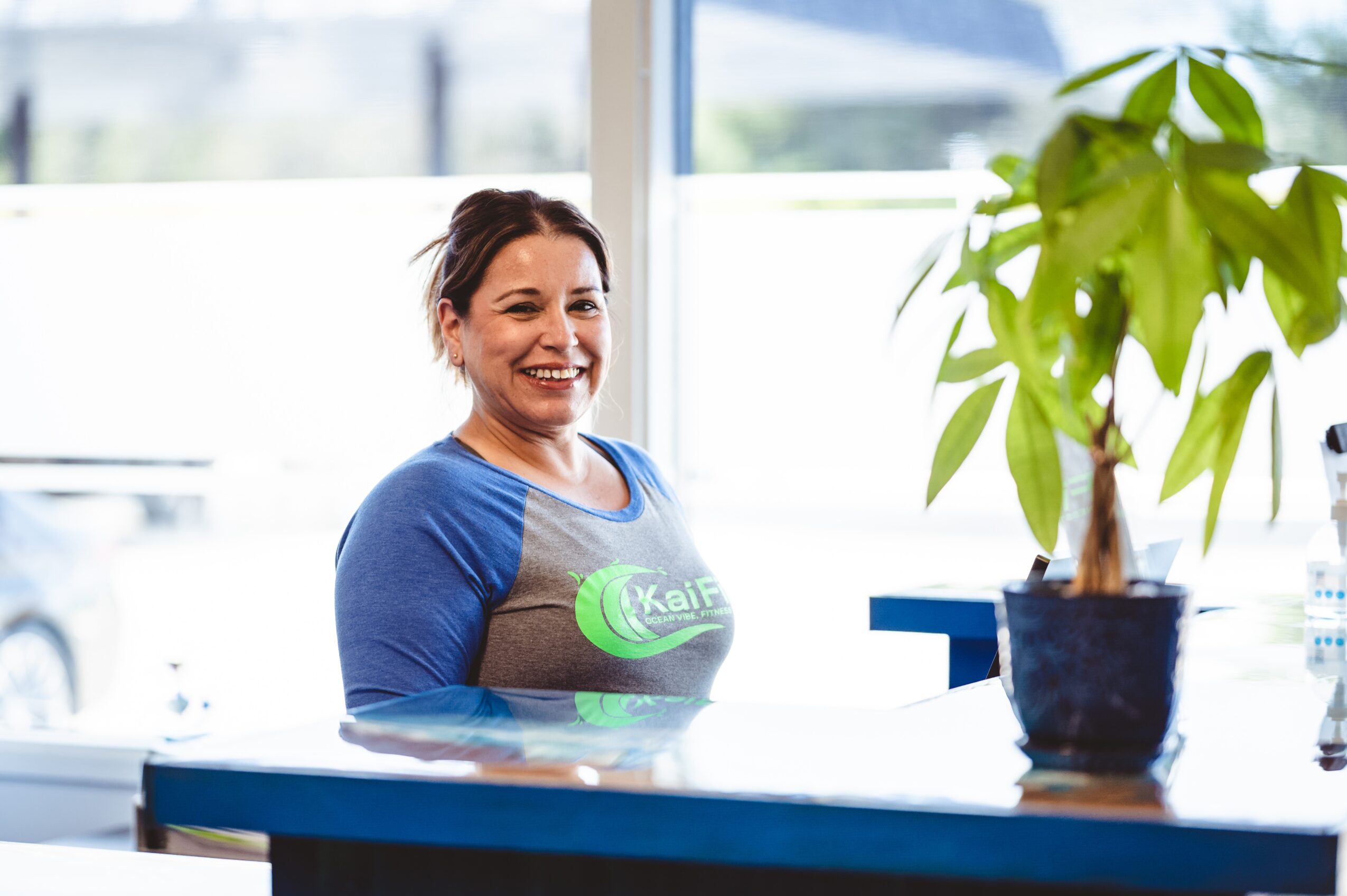Smiling woman with dark hair in a blue shirt standing indoors near a window, with a potted plant on a table.